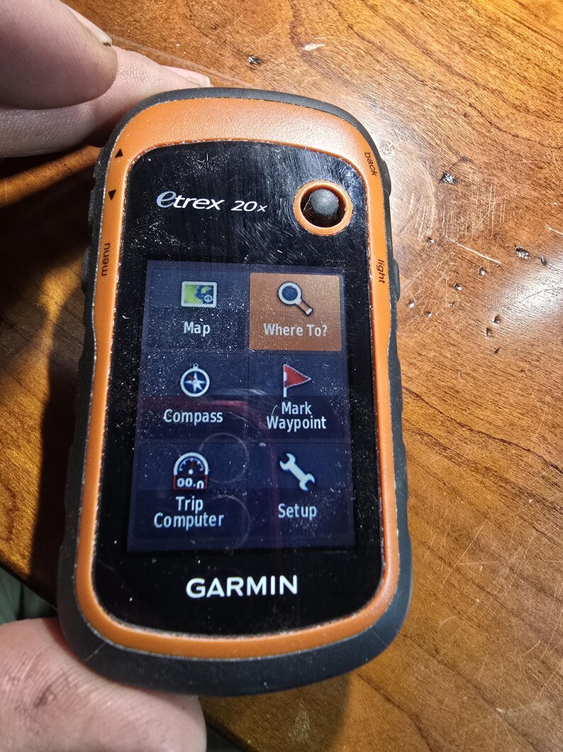 May include: A black and orange Garmin etrex 20x handheld GPS device with a color screen displaying a map, compass, and other navigation features. The screen also shows the text "Where To?" and "Mark Waypoint".