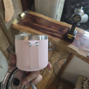 K&ouml;nnte beinhalten: Ein hellrosa Becher mit silbernem Rand und Griff. Der Becher wird in der Hand einer Person gehalten. Ein klarer Deckel ist sichtbar. Der Hintergrund umfasst eine Holzoberfl&auml;che und einen Akkuschrauber.