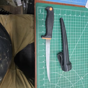 May include: A black and orange handled knife with a silver blade, approximately 12 inches long, next to a black sheath. The knife is on a green cutting mat with inch measurements.