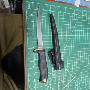 May include: A stainless steel fillet knife with a black handle and orange accents, next to a black sheath. The blade is approximately 7 inches long. The knife is on a green cutting mat.