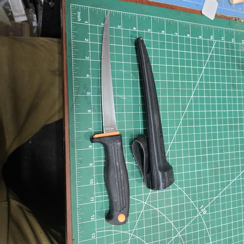 May include: A stainless steel fillet knife with a black handle and orange accents, next to a black sheath. The blade is approximately 7 inches long. The knife is on a green cutting mat.