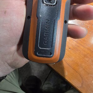 May include: A black and orange handheld GPS device with a silver loop on the top. The device has the word "GARMIN" printed on the side.