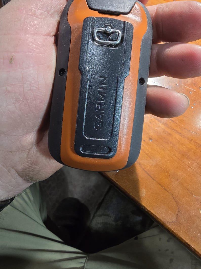 May include: A black and orange handheld GPS device with a silver loop on the top. The device has the word "GARMIN" printed on the side.
