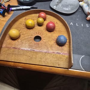 K&ouml;nnte beinhalten: Ein Vintage-Holzballspiel mit gew&ouml;lbter Oberseite und einer Reihe von L&ouml;chern. Das Spielbrett zeigt mehrere farbige Holzkugeln, darunter gelbe, rote und blaue. Das Brett ist mit Zahlen und dem Wort "Ball" markiert.