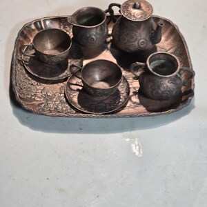 Op de afbeelding: Een miniatuur koperen theeservies met een theepot, roomkan, suikerpot en twee kopjes en schoteltjes. De set staat op een decoratief dienblad met bloemmotieven.