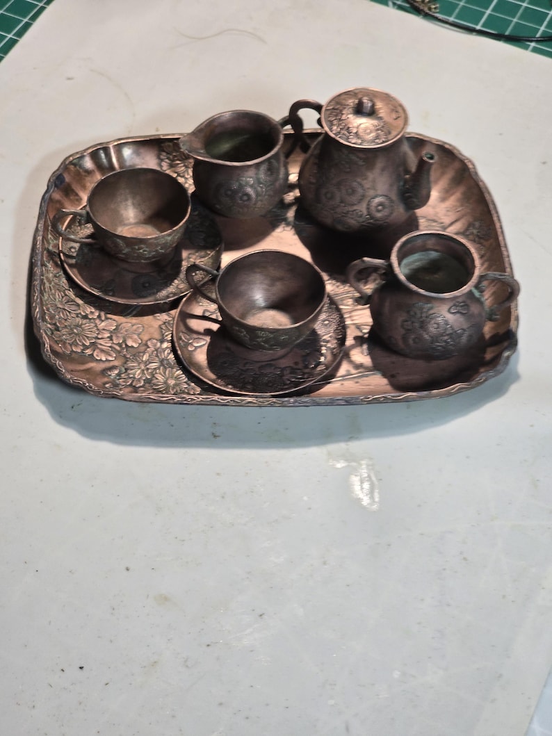 Op de afbeelding: Een miniatuur koperen theeservies met een theepot, roomkan, suikerpot en twee kopjes en schoteltjes. De set staat op een decoratief dienblad met bloemmotieven.