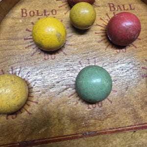 K&ouml;nnte beinhalten: Holzspielbrett mit den W&ouml;rtern "BOLLO" und "BALL" aufgedruckt. Das Brett zeigt mehrere farbige Holzkugeln: gelb, gr&uuml;n und rot. Das Brett hat ein abgenutztes, Vintage-Aussehen.