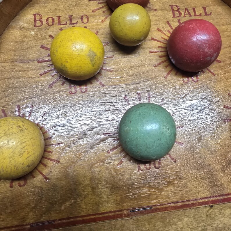 K&ouml;nnte beinhalten: Holzspielbrett mit den W&ouml;rtern "BOLLO" und "BALL" aufgedruckt. Das Brett zeigt mehrere farbige Holzkugeln: gelb, gr&uuml;n und rot. Das Brett hat ein abgenutztes, Vintage-Aussehen.