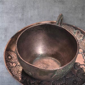 Op de afbeelding: Een vintage koperen theekopje en schotelset met een bloemenmotief. Het kopje en de schotel zijn beide versierd met een reli&euml;fbloemenpatroon.