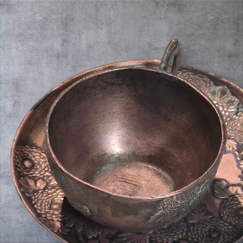 Op de afbeelding: Een vintage koperen theekopje en schotelset met een bloemenmotief. Het kopje en de schotel zijn beide versierd met een reli&euml;fbloemenpatroon.