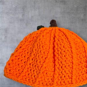 Puede incluir: Un gorro de calabaza tejido a crochet de color naranja brillante. El gorro tiene una superficie texturizada y un tallo en la parte superior.