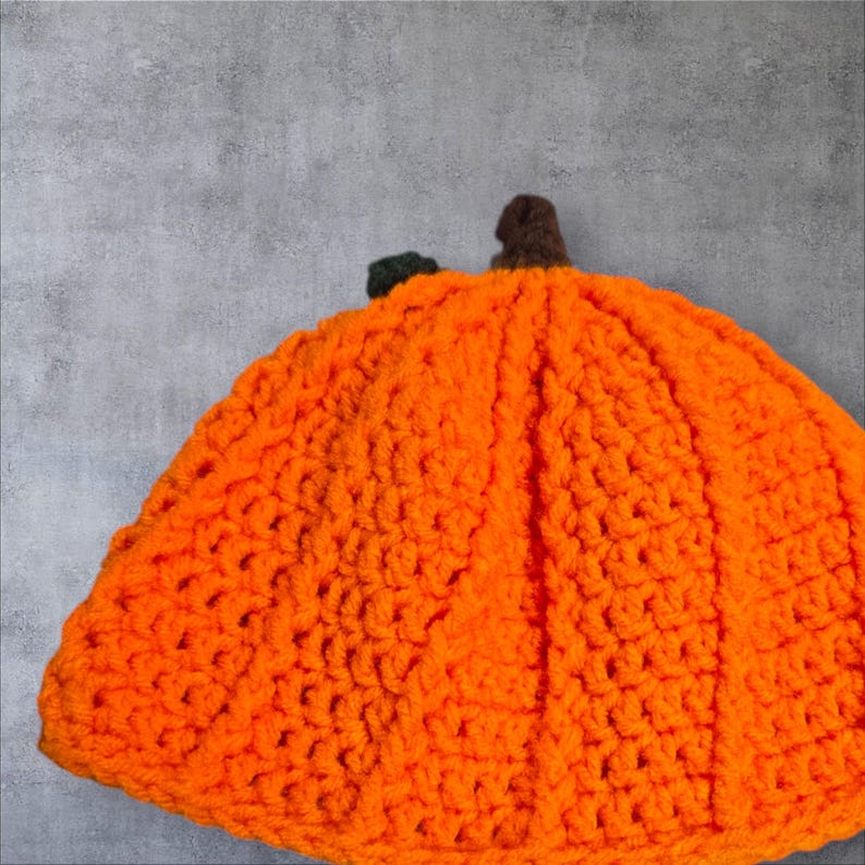 Puede incluir: Un gorro de calabaza tejido a crochet de color naranja brillante. El gorro tiene una superficie texturizada y un tallo en la parte superior.
