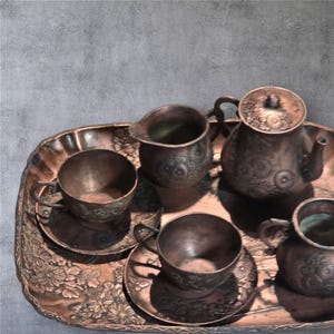 Op de afbeelding: Een koperen theeset op een decoratief dienblad. De set bevat een theepot, een roomkan, een suikerpot en twee theekopjes met schoteltjes.