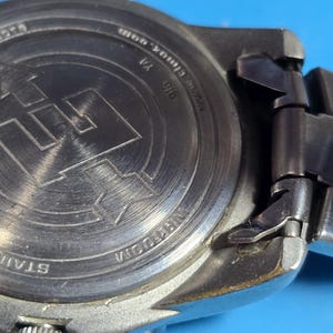 May include: The back of a silver watch with a stainless steel band. The watch face has a black background with white markings. The watch has a date window at the 3 o'clock position. The watch is water resistant and has a screw down crown. The watch has a black leather band.