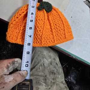Puede incluir: Sombrero de calabaza de punto de ganchillo naranja con hojas y tallo de fieltro verde. El sombrero se est&aacute; midiendo con una cinta m&eacute;trica, mostrando que tiene aproximadamente 18 cm de alto.