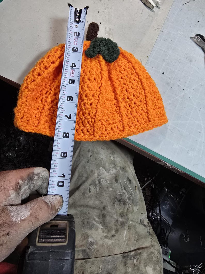 Puede incluir: Sombrero de calabaza de punto de ganchillo naranja con hojas y tallo de fieltro verde. El sombrero se est&aacute; midiendo con una cinta m&eacute;trica, mostrando que tiene aproximadamente 18 cm de alto.
