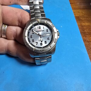 May include: A silver Timex Expedition watch with a white face and black numbers. The watch has a date window and a day of the week window. The watch is on a silver metal band.