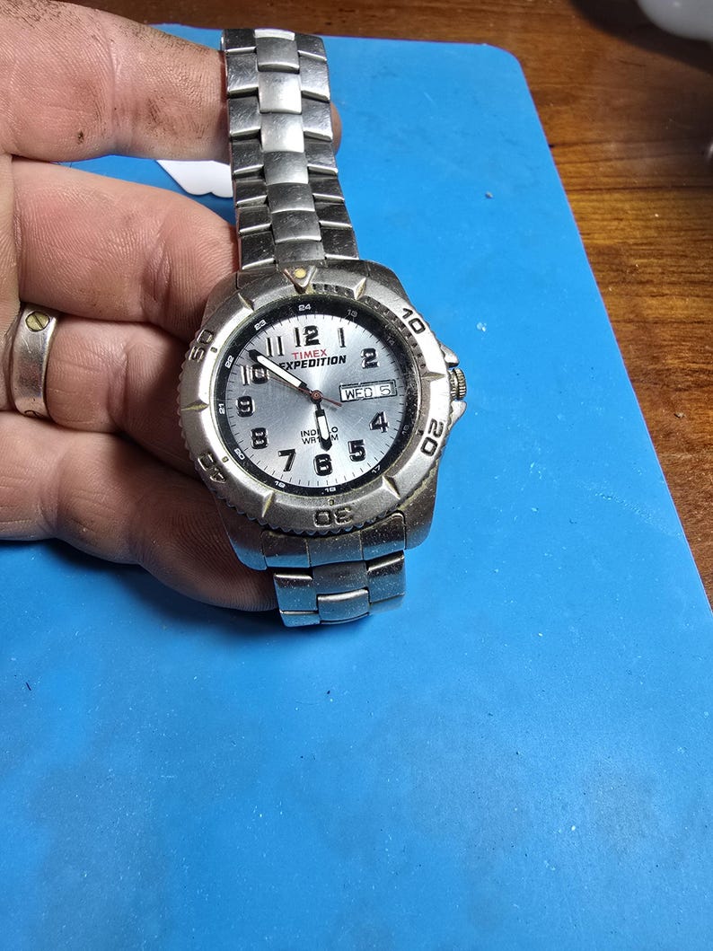 May include: A silver Timex Expedition watch with a white face and black numbers. The watch has a date window and a day of the week window. The watch is on a silver metal band.
