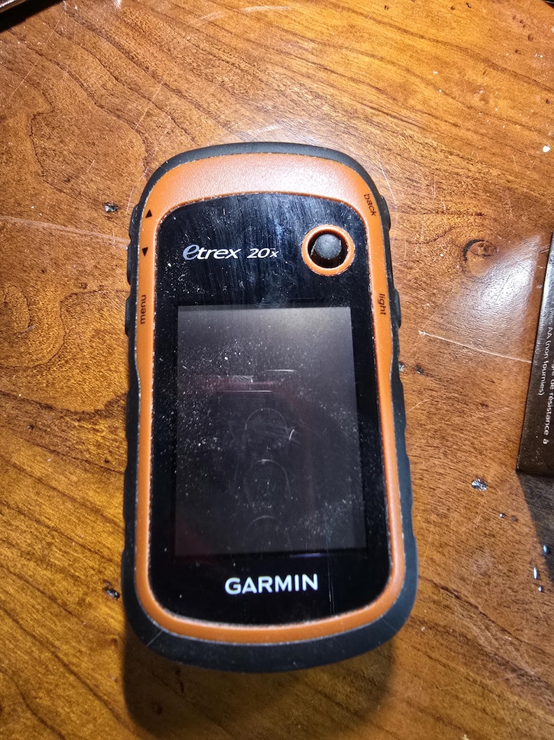 May include: A black and orange Garmin eTrex 20x handheld GPS device with a screen that is turned off. The device has a black and orange casing with a black button on the top right corner. The device is sitting on a wooden surface.