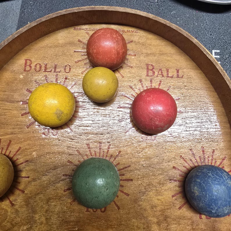 K&ouml;nnte beinhalten: Holz-Bollo-Ball-Spielbrett mit sechs bunten Kugeln. Auf dem Holzbrett steht "Bollo Ball". Die Kugeln sind rot, gelb, gr&uuml;n und blau.