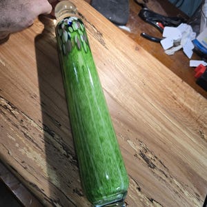 May include: A green glass rolling pin with clear glass ends. The rolling pin has a mottled green and white design with blue and brown accents.