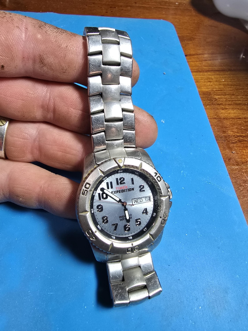 May include: A silver Timex Expedition watch with a black face and silver hands. The watch has a date window that says "WED 51".