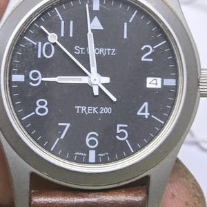 May include: A St. Moritz Trek 200 wristwatch with a round silver-tone metal case and a black dial. The dial features white Arabic numerals, a date window at the three o'clock position, a triangle marker at twelve, and the text Japan Mov't.