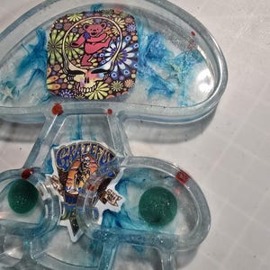 May include: A clear resin mushroom-shaped trinket with a blue and white swirl design. The mushroom has two green balls inside the cap and a Grateful Dead sticker on the stem. The sticker features a skull and crossbones with the words "Grateful Dead" and "Since 1965".