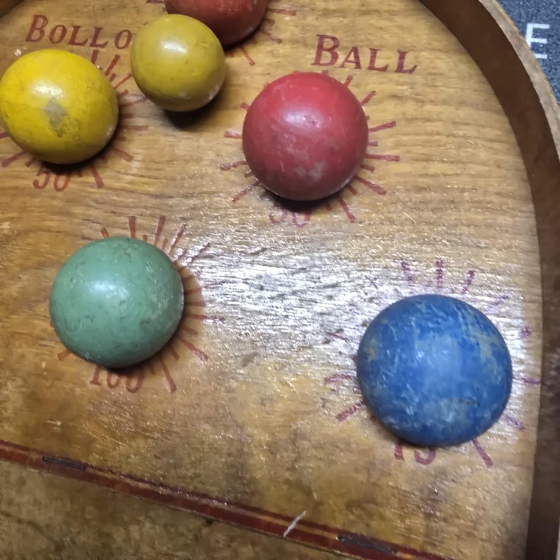 K&ouml;nnte beinhalten: Ein h&ouml;lzernes Bollo Ball-Spielbrett mit bunten Kugeln. Auf dem Brett stehen die Worte "BALL" und "BOLLO". Die Kugeln sind gelb, rot, gr&uuml;n und blau.