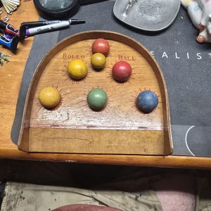 K&ouml;nnte beinhalten: Ein Vintage-Bollo-Ball-Spiel mit Holzrahmen und bunten Kugeln. Die Kugeln sind gelb, rot, gr&uuml;n und blau. Auf dem Spielbrett steht "Bollo Ball".