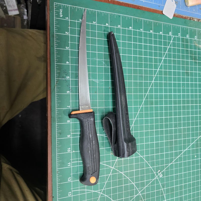 May include: A stainless steel fillet knife with a black handle and orange accents, next to its black sheath. The blade is approximately 7 inches long. The knife is on a green cutting mat.