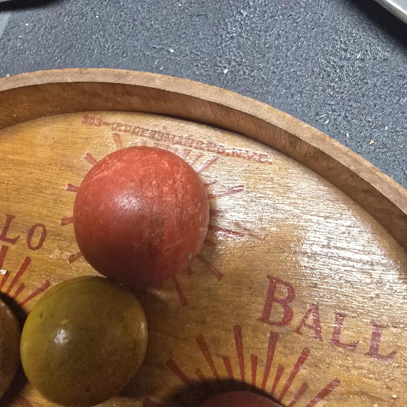 K&ouml;nnte beinhalten: Holzspielbrett mit einer roten Kugel und einer gr&uuml;nen Kugel. Das Brett hat das Wort "BALL" in roten Buchstaben und ein Sonnenstrahl-Design. Der Text "303-O.PRESBMAR226.N.Y.C." ist auf dem Brett aufgedruckt.