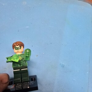 May include: A Green Lantern Lego minifigure with a green and black costume, brown hair, and a green mask. The figure is holding a green translucent accessory. The base reads "WORLD MINIFIGURES Collect Them All."