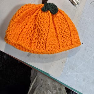 Puede incluir: Un gorro de calabaza de ganchillo de color naranja con un tallo y una hoja verdes. El gorro est&aacute; sobre una estera de corte verde.