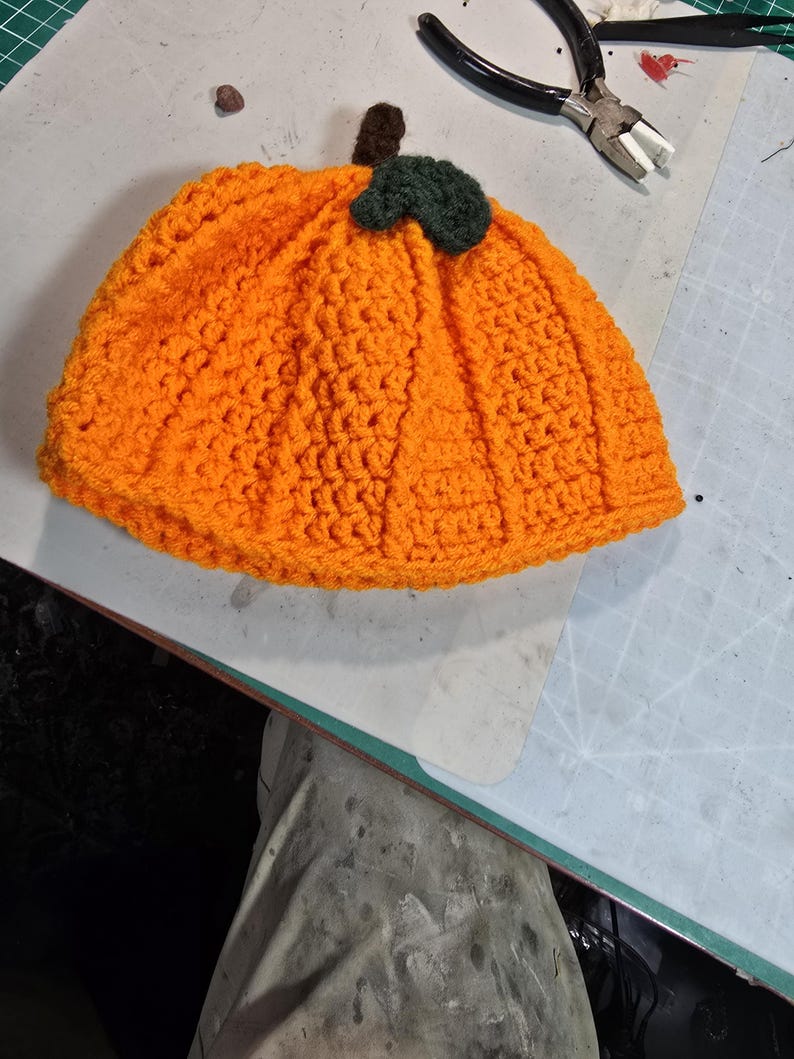 Puede incluir: Un gorro de calabaza de ganchillo de color naranja con un tallo y una hoja verdes. El gorro est&aacute; sobre una estera de corte verde.