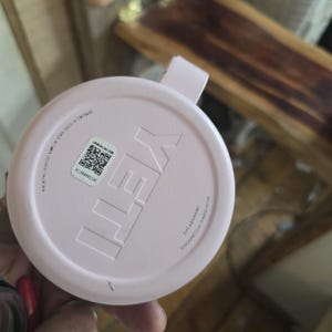 K&ouml;nnte beinhalten: Ein hellrosa YETI-Deckel mit einem kleinen Griff. Der Deckel hat das YETI-Logo in einer erhabenen, helleren rosa Farbe. Der Deckel hat auch einen Aufkleber mit einem QR-Code und Text. Der Deckel besteht aus einem strapazierf&auml;higen Material.