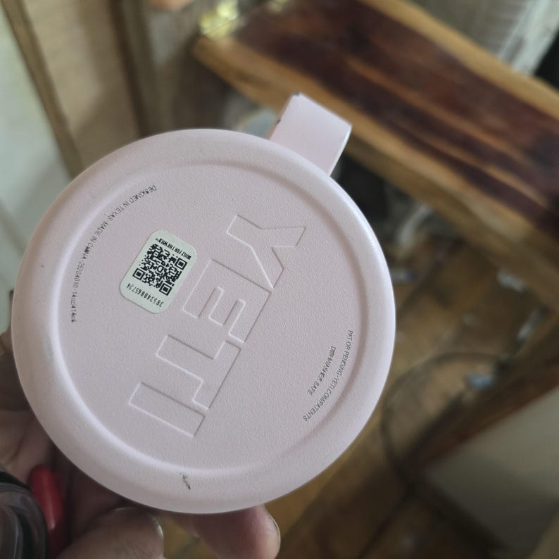 K&ouml;nnte beinhalten: Ein hellrosa YETI-Deckel mit einem kleinen Griff. Der Deckel hat das YETI-Logo in einer erhabenen, helleren rosa Farbe. Der Deckel hat auch einen Aufkleber mit einem QR-Code und Text. Der Deckel besteht aus einem strapazierf&auml;higen Material.