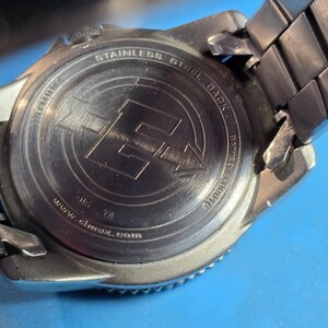 May include: Close-up of the back of a silver watch with a stainless steel black band. The watch face has the words "Stainless Steel Black" and "www.almax.co.uk" engraved on it.