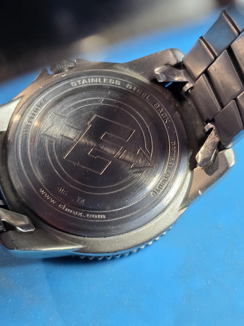 May include: Close-up of the back of a silver watch with a stainless steel black band. The watch face has the words "Stainless Steel Black" and "www.almax.co.uk" engraved on it.