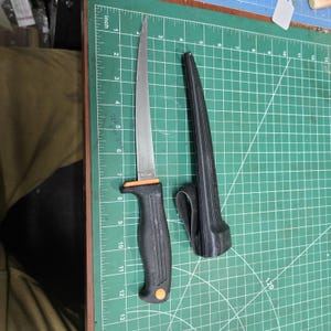 May include: A stainless steel fillet knife with a black handle and orange accents, next to its black sheath. The knife blade is approximately 7 inches long. The knife and sheath are on a green cutting mat.