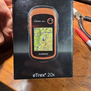 May include: A black and orange Garmin eTrex 20x GPS device with a map of Portland, Oregon displayed on the screen. The device is in its box, which is black with white text.