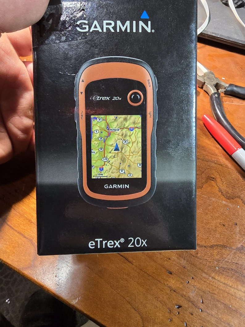 May include: A black and orange Garmin eTrex 20x GPS device with a map of Portland, Oregon displayed on the screen. The device is in its box, which is black with white text.