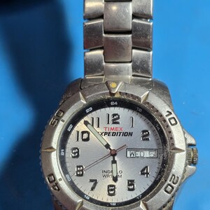 May include: A silver Timex Expedition watch with a white face and black hands. The watch has a date window and a day of the week window. The watch is water resistant to 100 meters.