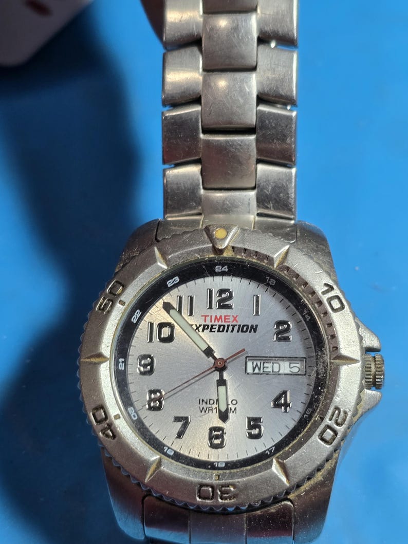 May include: A silver Timex Expedition watch with a white face and black hands. The watch has a date window and a day of the week window. The watch is water resistant to 100 meters.