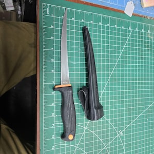 May include: A stainless steel fillet knife with a black handle and orange accents, next to its black sheath. The knife blade is approximately 8 inches long. The knife is on a green cutting mat.