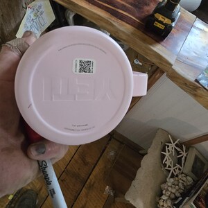 K&ouml;nnte beinhalten: Ein hellrosa Yeti-Becher mit Henkel. Der Markenname "YETI" ist auf der Unterseite des Bechers eingepr&auml;gt. Ein QR-Code und anderer Text sind auf einem Aufkleber zu sehen. Der Becher wird von einer Person gehalten.