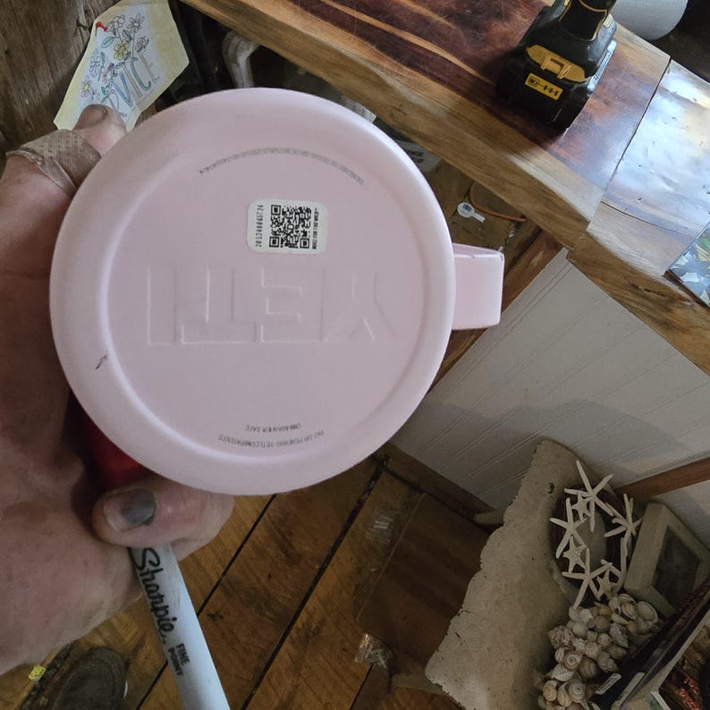 K&ouml;nnte beinhalten: Ein hellrosa Yeti-Becher mit Henkel. Der Markenname "YETI" ist auf der Unterseite des Bechers eingepr&auml;gt. Ein QR-Code und anderer Text sind auf einem Aufkleber zu sehen. Der Becher wird von einer Person gehalten.