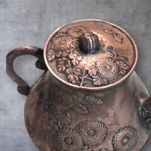 Op de afbeelding: Een koperen theepot met een deksel met een reli&euml;fmotief van chrysanten en bladeren. De theepot heeft een rond lichaam en een enkele handgreep.