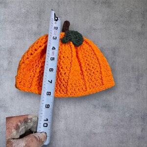 Puede incluir: Sombrero de calabaza de punto de ganchillo naranja con un tallo y una hoja verdes. El sombrero mide aproximadamente 15 cm de alto.