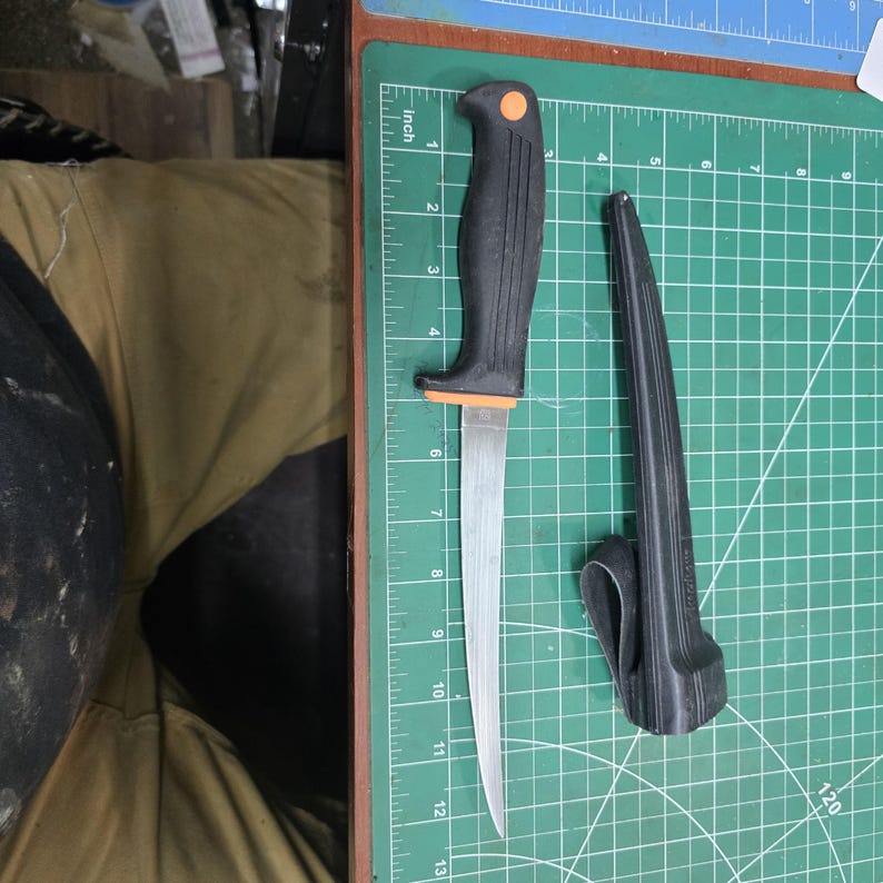 May include: A black-handled fillet knife with an orange accent and a silver blade, next to a black sheath. The knife is on a green cutting mat with inch measurements.